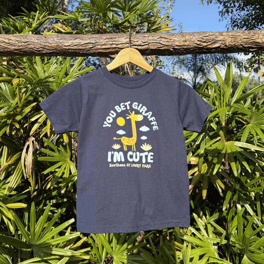A navy blue kids' t-shirt with a yellow and white giraffe graphic hanging on a wooden hanger from a tree branch outdoors, surrounded by green palm leaves.