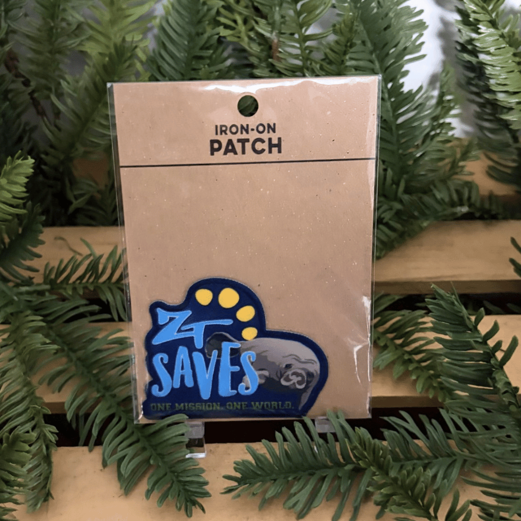 An iron-on patch in its clear plastic packaging, displayed on a wooden shelf surrounded by green pine branches. The patch features a blue and yellow design with the text 'ZT SAVES' and 'ONE MISSION, ONE WORLD' with a manatee.