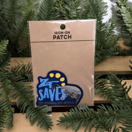 An iron-on patch in its clear plastic packaging, displayed on a wooden shelf surrounded by green pine branches. The patch features a blue and yellow design with the text 'ZT SAVES' and 'ONE MISSION, ONE WORLD' with a manatee.