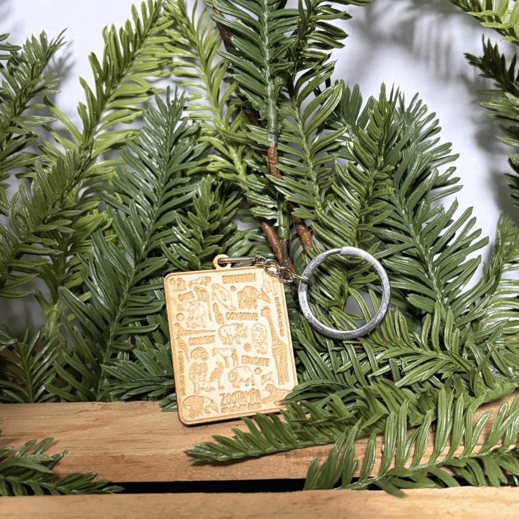 A rectangular engraved wooden charm keychain with a silver metal key ring, displayed on green pine branches over a wooden surface.