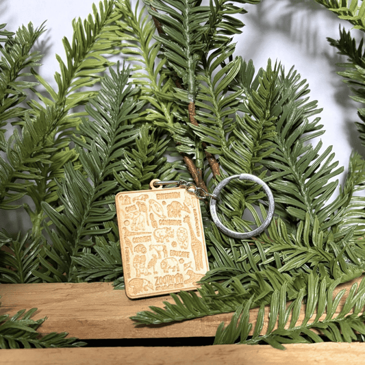A rectangular engraved wooden charm keychain with a silver metal key ring, displayed on green pine branches over a wooden surface.