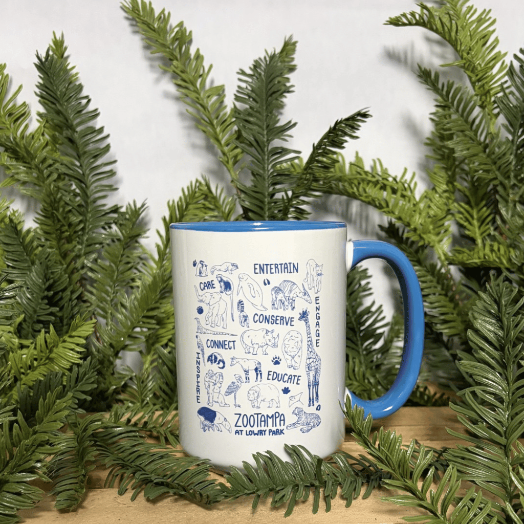 A white ceramic mug with a blue handle and blue interior, decorated with blue line-art illustrations and text from ZooTampa, including 'Entertain', 'Conserve', 'Connect', 'Educate', and 'ZooTampa at Lowry Park'. The mug is styled on a wooden surface surrounded by green pine branches against a light gray background.