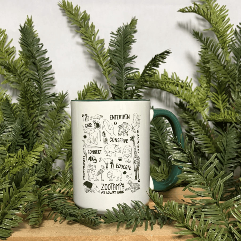 A white ceramic mug with a dark green handle and interior, featuring black hand-drawn illustrations and text including 'ZooTampa at Lowry Park,' styled with green pine branches on a wooden surface against a white background.