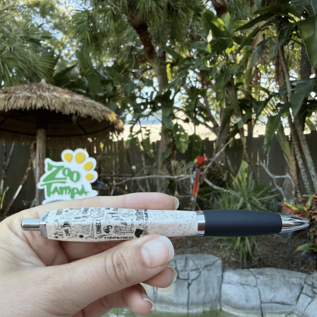 A hand holds a pen with a beige speckled barrel featuring black cartoon-style line art and a black grip, with a Zoo Tampa sign blurred in the background outdoors.