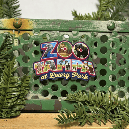 A colorful magnet shaped like a zoo map with the text 'ZooTampa at Lowry Park,' displayed on a distressed green metal crate with pine branches around it.