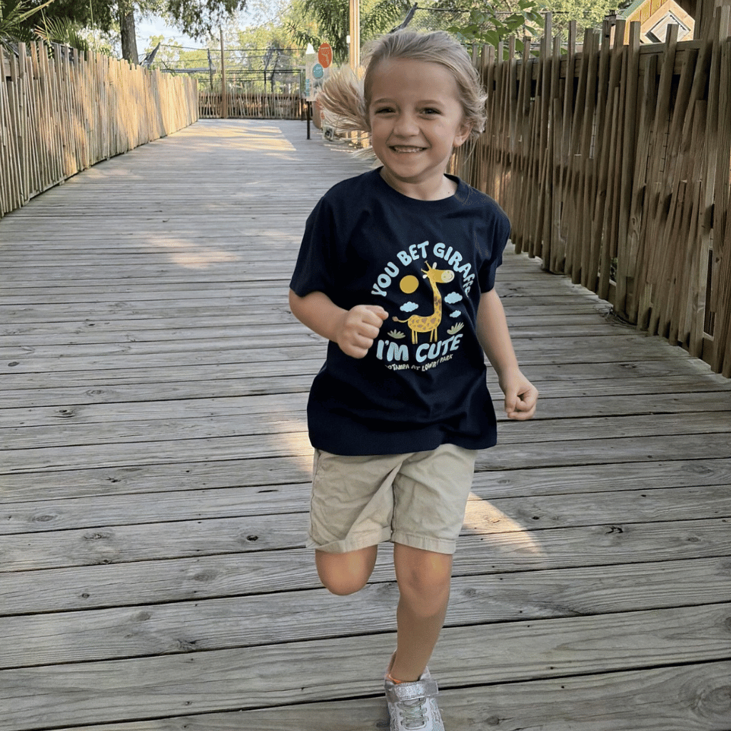 A young child running on a wooden boardwalk wearing a navy blue graphic t-shirt with a giraffe and the text 'You Bet Giraffe I'm Cute', paired with khaki shorts and white sneakers.