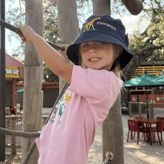 A young child wearing a pink t-shirt and a navy blue 'ZooTampa' bucket hat while climbing on an outdoor playground with wooden posts and ropes.