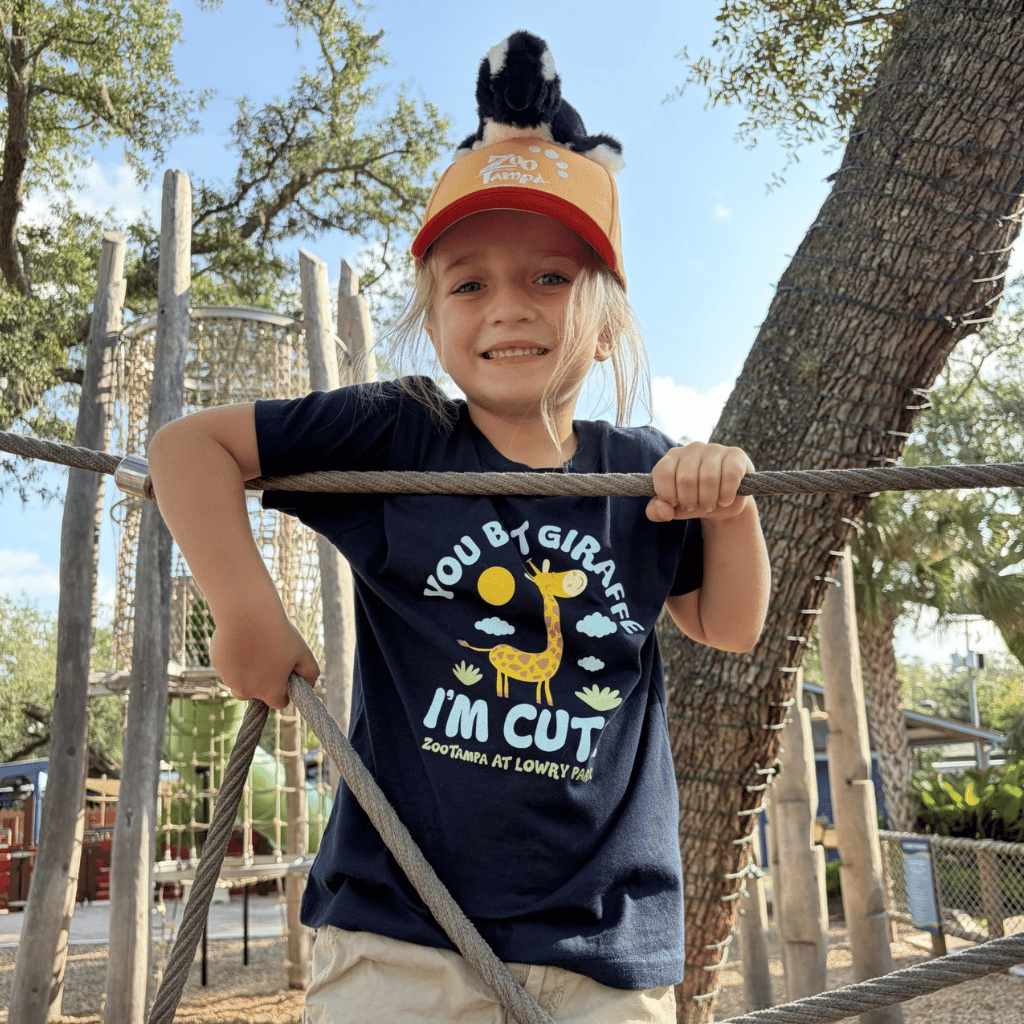 A young girl smiling at a playground, wearing a navy blue graphic t-shirt that reads 'You Bet Giraffe I'm Cute' with a giraffe illustration, and a bright orange cap with a small black-and-white penguin sitting on top.