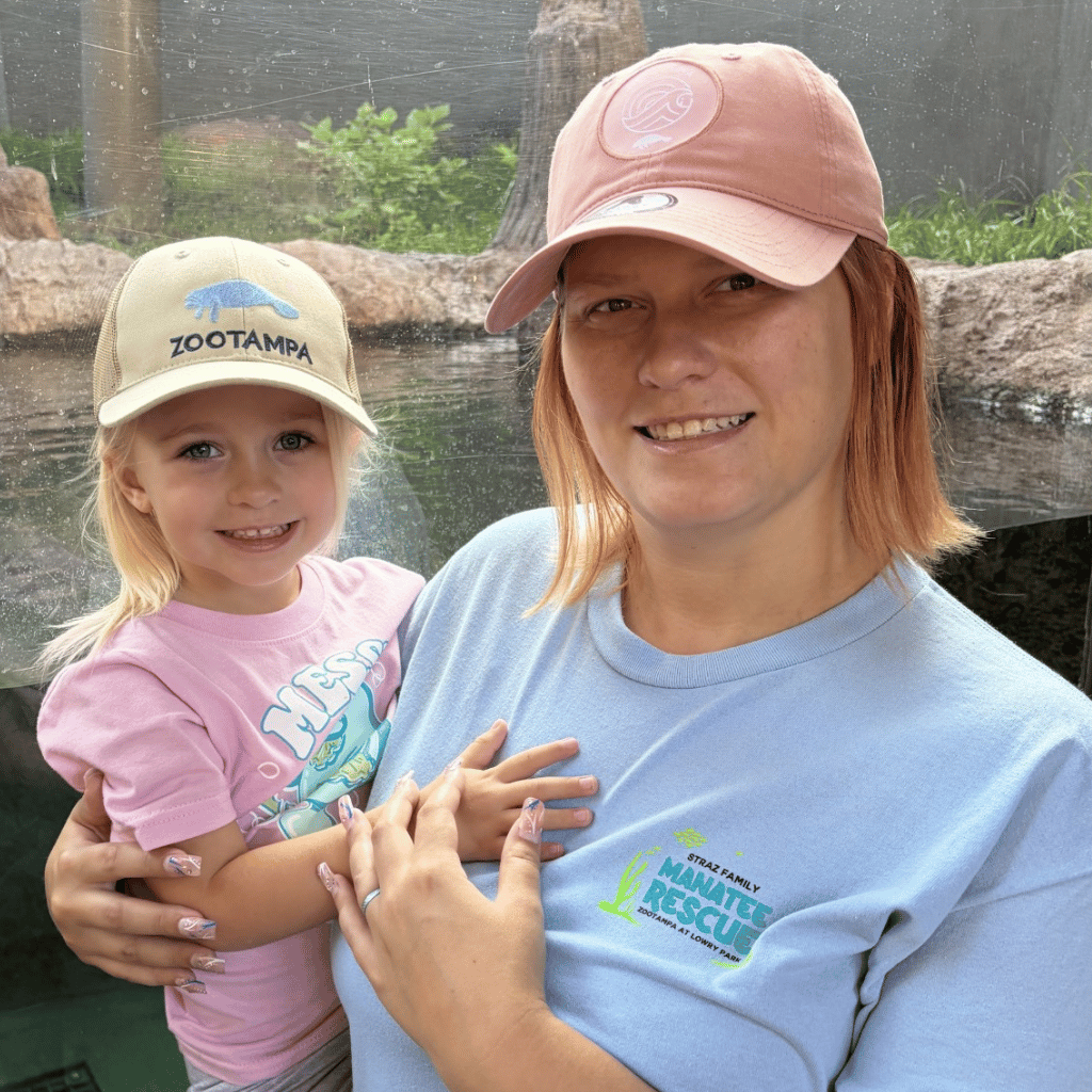 A woman and a young girl smiling while wearing matching pink and beige Zootampa t-shirts and baseball caps, standing in front of a zoo enclosure.