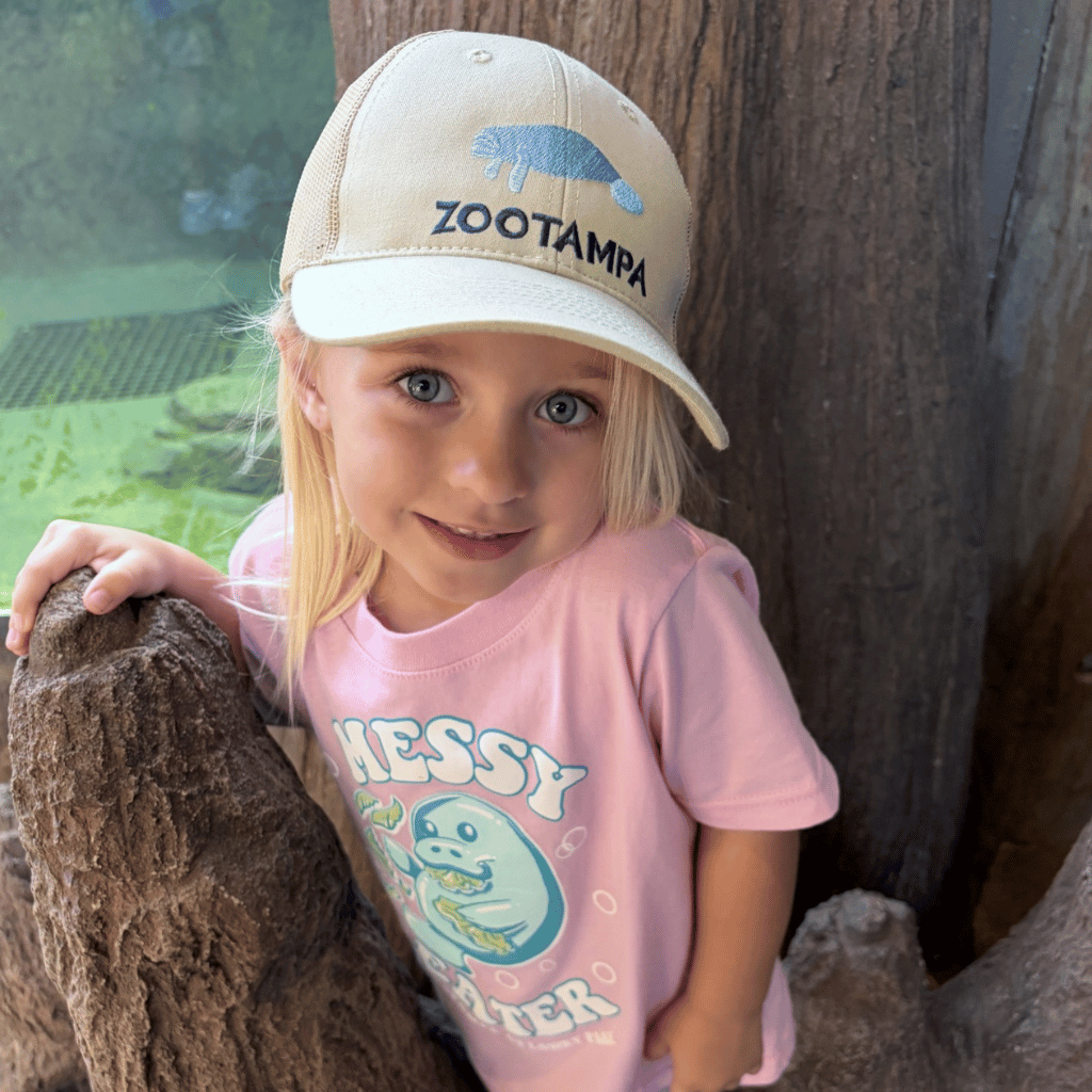A young girl with blonde hair wearing a beige Zootampa baseball cap and a pink graphic t-shirt, posing outdoors next to a tree trunk.