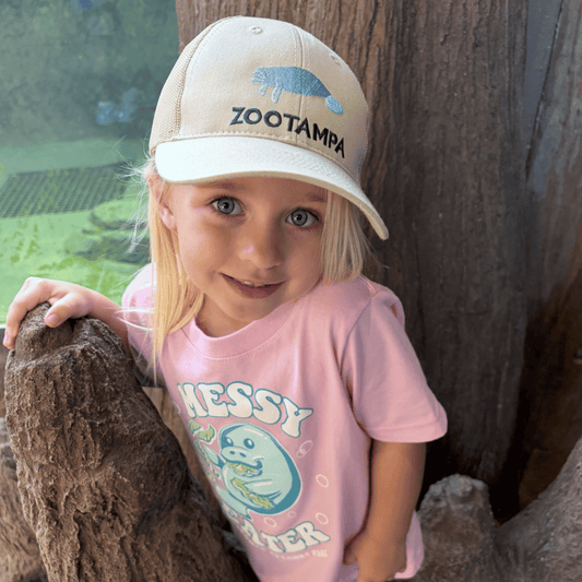 A young girl with blonde hair wearing a beige Zootampa baseball cap and a pink graphic t-shirt, posing outdoors next to a tree trunk.