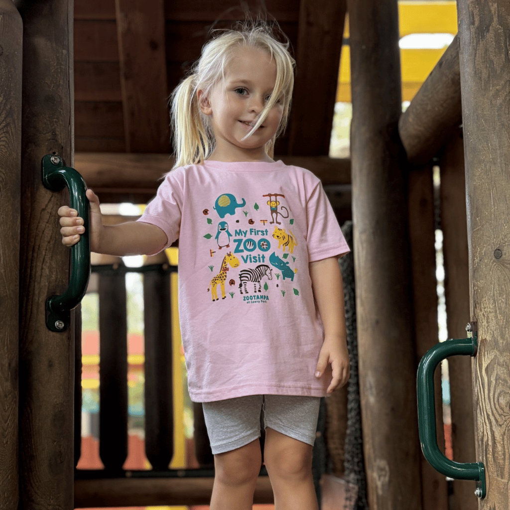 A young blonde girl wearing a light pink graphic t-shirt that reads 'My First Zoo Visit' with cartoon animals, standing on a wooden playground structure.
