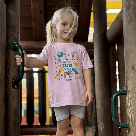 A young blonde girl wearing a light pink graphic t-shirt that reads 'My First Zoo Visit' with cartoon animals, standing on a wooden playground structure.