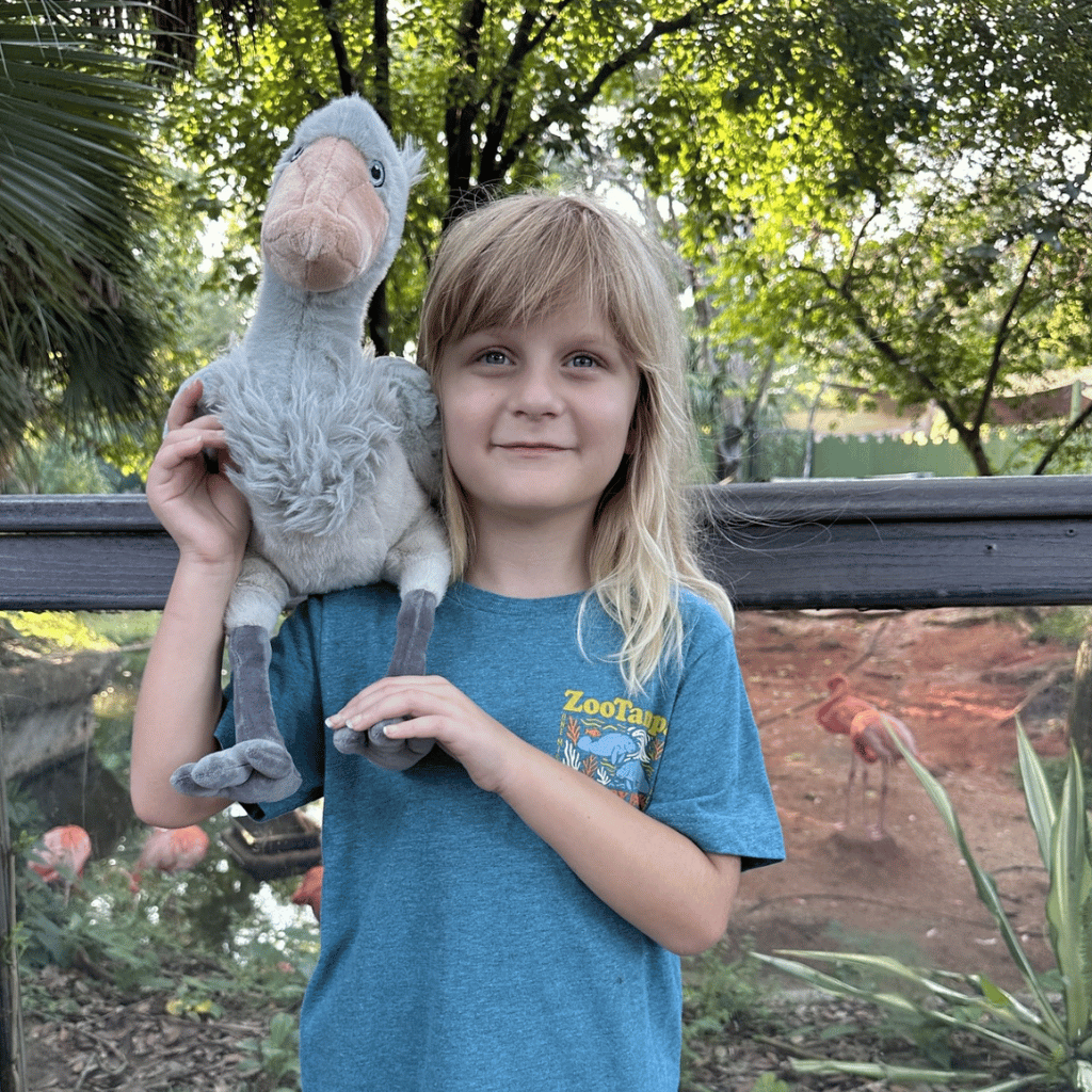 A young girl with blonde hair wearing a blue t-shirt holds a shoebill stork plush toy. The background shows a zoo habitat with trees, a fence, and flamingos.