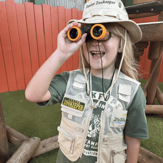 A young child wearing a beige safari-style zookeeper hat and matching vest over an olive green shirt, holding orange binoculars to their eyes in an outdoor play area with a red wooden fence in the background.