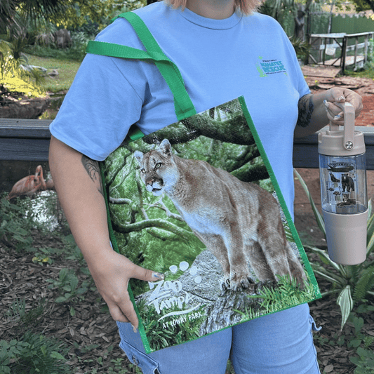 A person outdoors wearing a light blue T-shirt and jeans, holding a large printed tote bag with a cougar graphic and green straps, while also holding a clear water bottle with a beige cap.